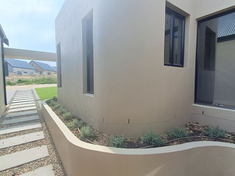 A modern exterior view featuring a curved retaining wall, landscaped garden bed with irrigation, and a stone pathway leading along the side of the house.