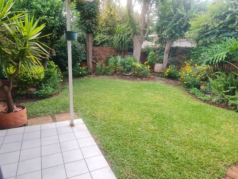 A private, covered patio area overlooking a well-maintained, lush green garden with established trees and a brick boundary wall.
