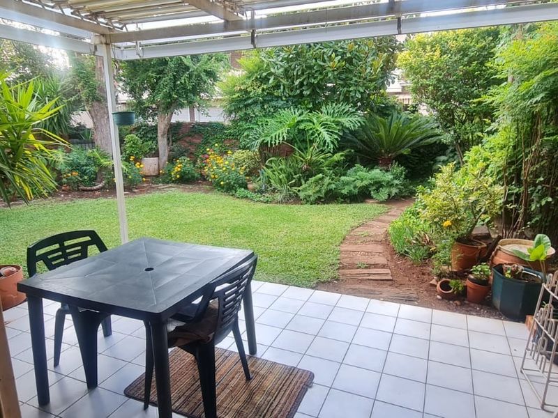 A covered patio area featuring tiled flooring, overlooking a lush, established garden with a brick boundary wall and a paved pathway.