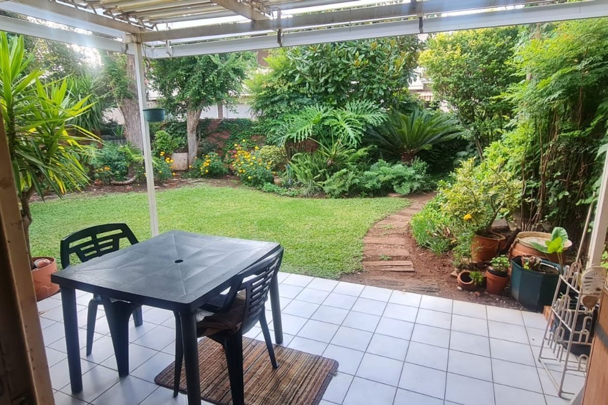 A covered patio area featuring tiled flooring, overlooking a lush, established garden with a brick boundary wall and a paved pathway.