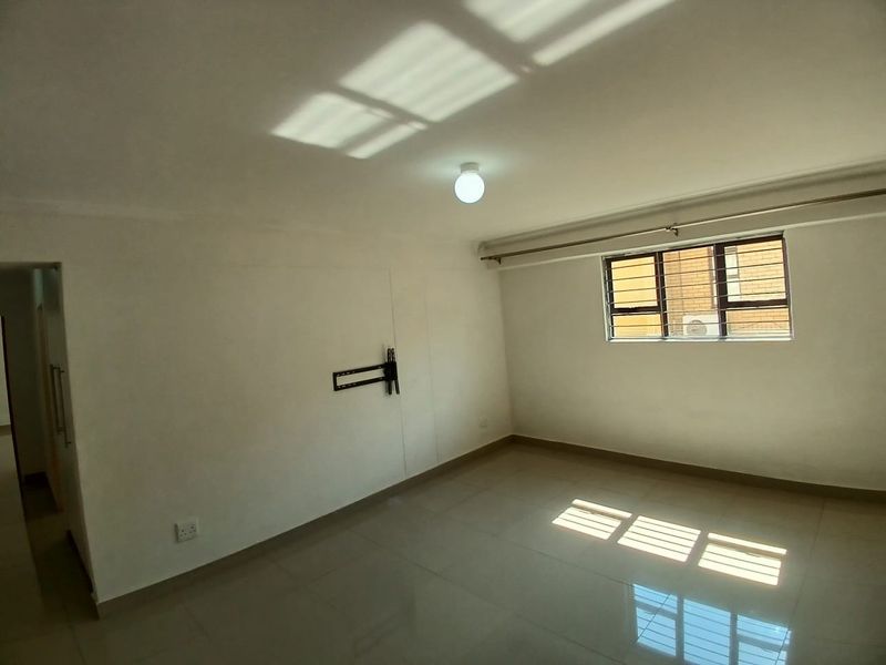 A bright, empty living room featuring tiled flooring, a wall-mounted television bracket, and a window with security bars.