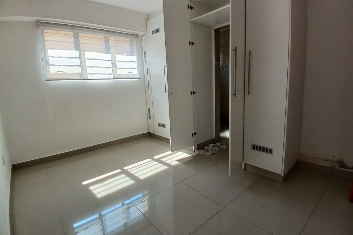 A bright, empty bedroom featuring tiled flooring, a roller blind, and built-in white cupboards with ample storage space.