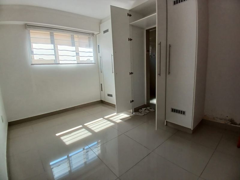 A bright, empty bedroom featuring tiled flooring, a roller blind, and built-in white cupboards with ample storage space.