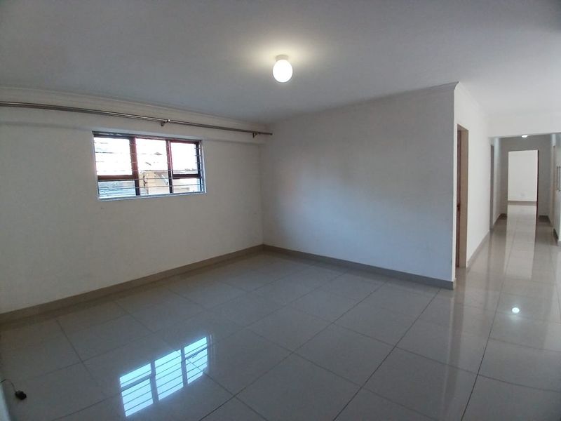 A spacious, empty living area featuring neutral tiled flooring, clean white walls, and a window fitted with security bars.