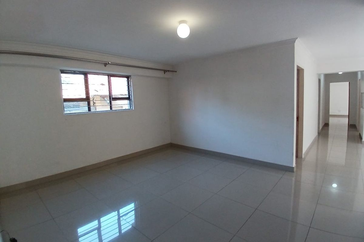 A spacious, empty living area featuring neutral tiled flooring, clean white walls, and a window fitted with security bars.