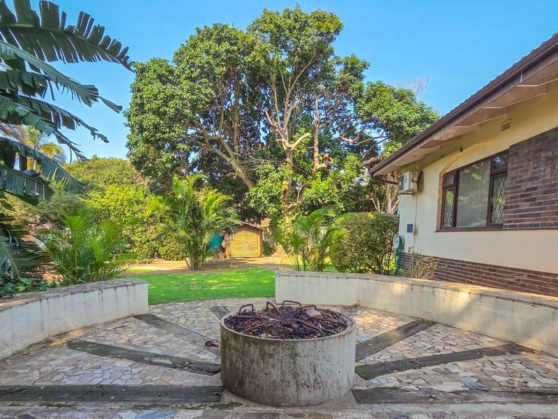 Rear exterior view featuring a paved patio area with a built-in fire pit, overlooking a spacious garden with mature trees.