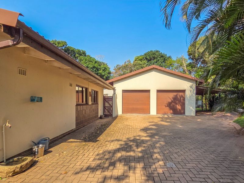 A view of the rear exterior featuring a paved driveway leading to a double garage with roll-up doors and surrounding garden area.