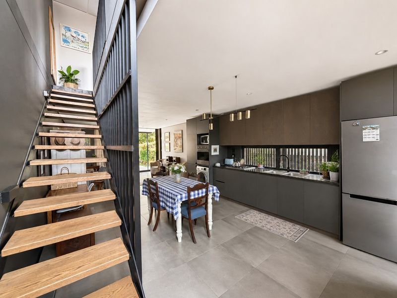 Modern open-plan kitchen and dining area featuring sleek dark cabinetry, integrated appliances, tiled flooring, and a striking timber staircase with metal balustrade.