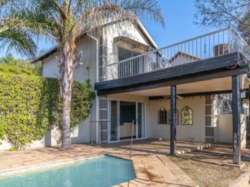 Rear exterior view featuring a swimming pool, paved deck, covered patio area, and an upper-level balcony with metal railings.