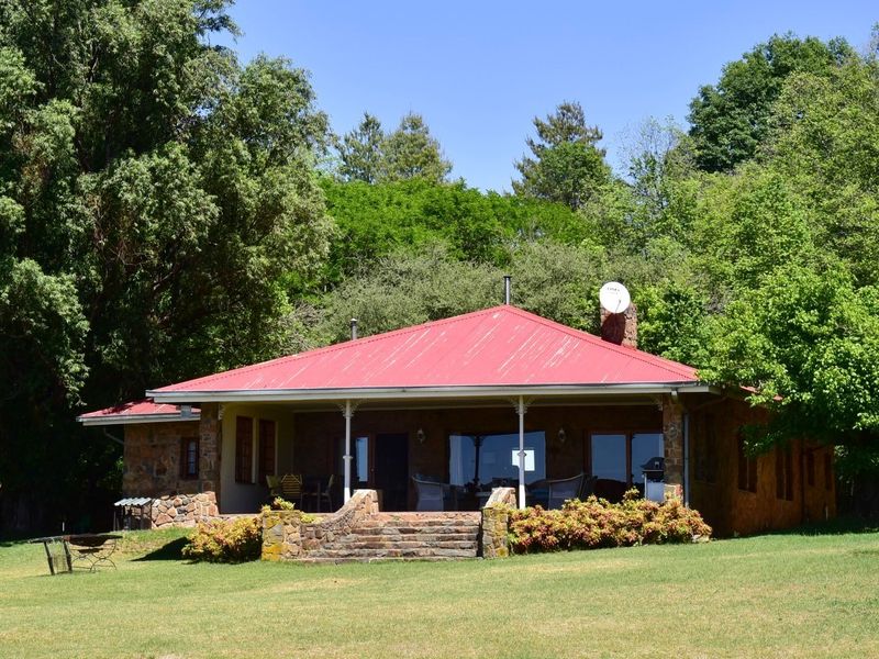 A charming farmhouse exterior featuring stone masonry walls, a red corrugated metal roof, and a spacious covered patio overlooking a lush garden.
