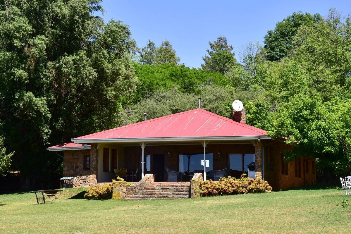 A charming farmhouse exterior featuring stone masonry walls, a red corrugated metal roof, and a spacious covered patio overlooking a lush garden.