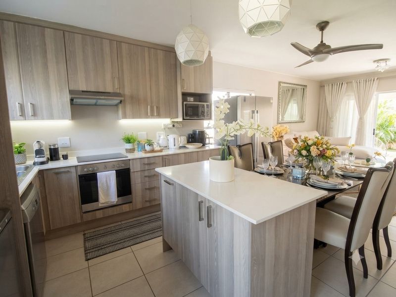 Modern open-plan kitchen and dining area featuring light wood cabinetry, stone countertops, a central island, and integrated appliances with tiled flooring throughout.