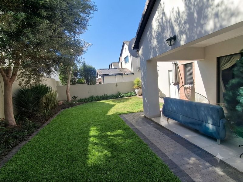 A well-maintained covered patio area featuring paved flooring, overlooking a neat, landscaped garden with a lawn and boundary wall.