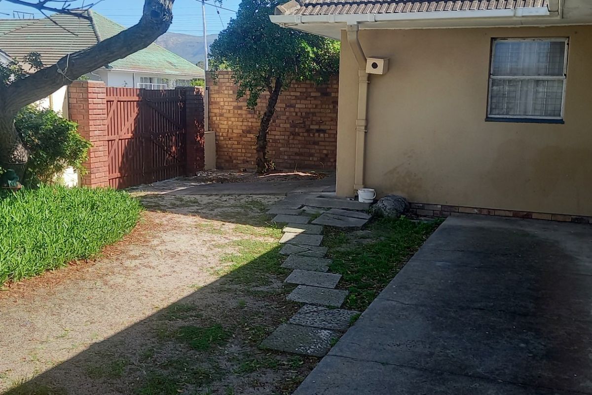 A rear exterior view of the property featuring a concrete pathway, brick boundary wall, and a small garden area with mature trees.