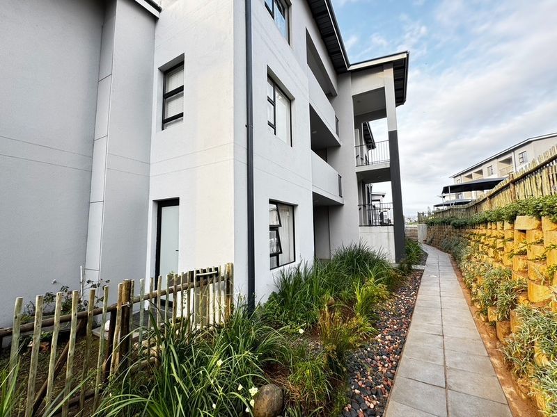 Exterior view of the apartment building featuring a paved walkway, landscaped garden beds, and private balconies with metal railings.