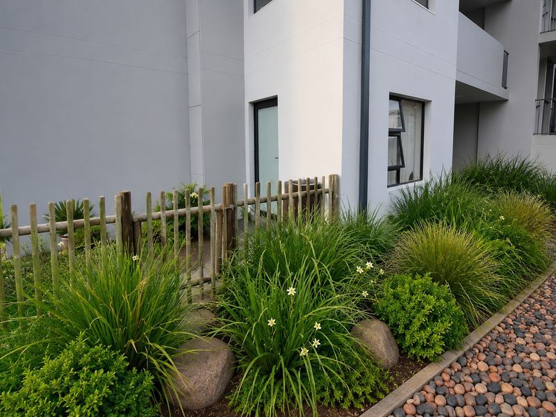 A well-maintained rear exterior view of the apartment featuring a timber picket fence, landscaped garden beds with pebble borders, and modern building finishes.