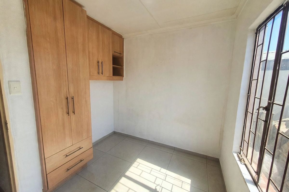 A bright, empty bedroom featuring light wood-finish built-in cupboards, neutral floor tiles, and security bars on the window.