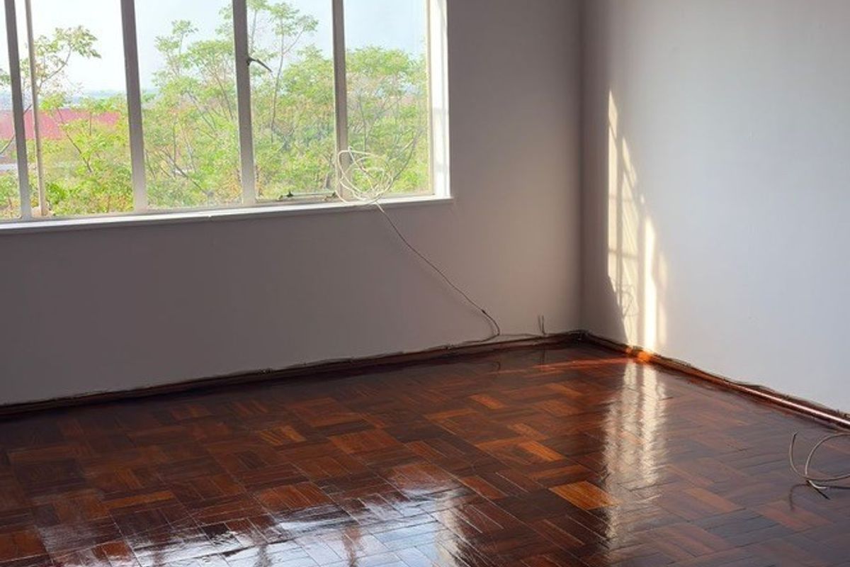 A bright, empty bedroom featuring polished parquet flooring, a large window with natural light, and simple white walls.