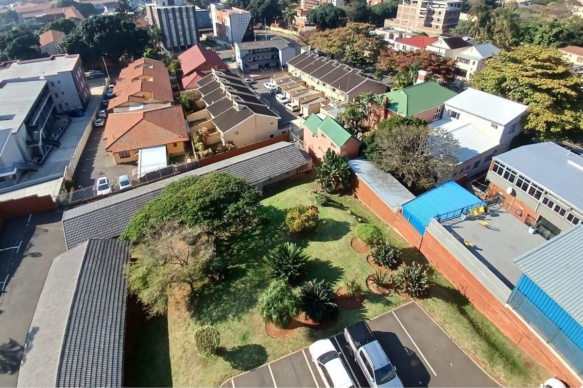 An aerial view of the apartment complex grounds, featuring a central garden area, paved parking bays, and surrounding residential buildings.