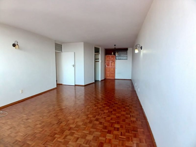 A spacious, empty living area featuring classic parquet flooring, neutral walls, and wall-mounted lighting, providing a versatile blank canvas for interior design.