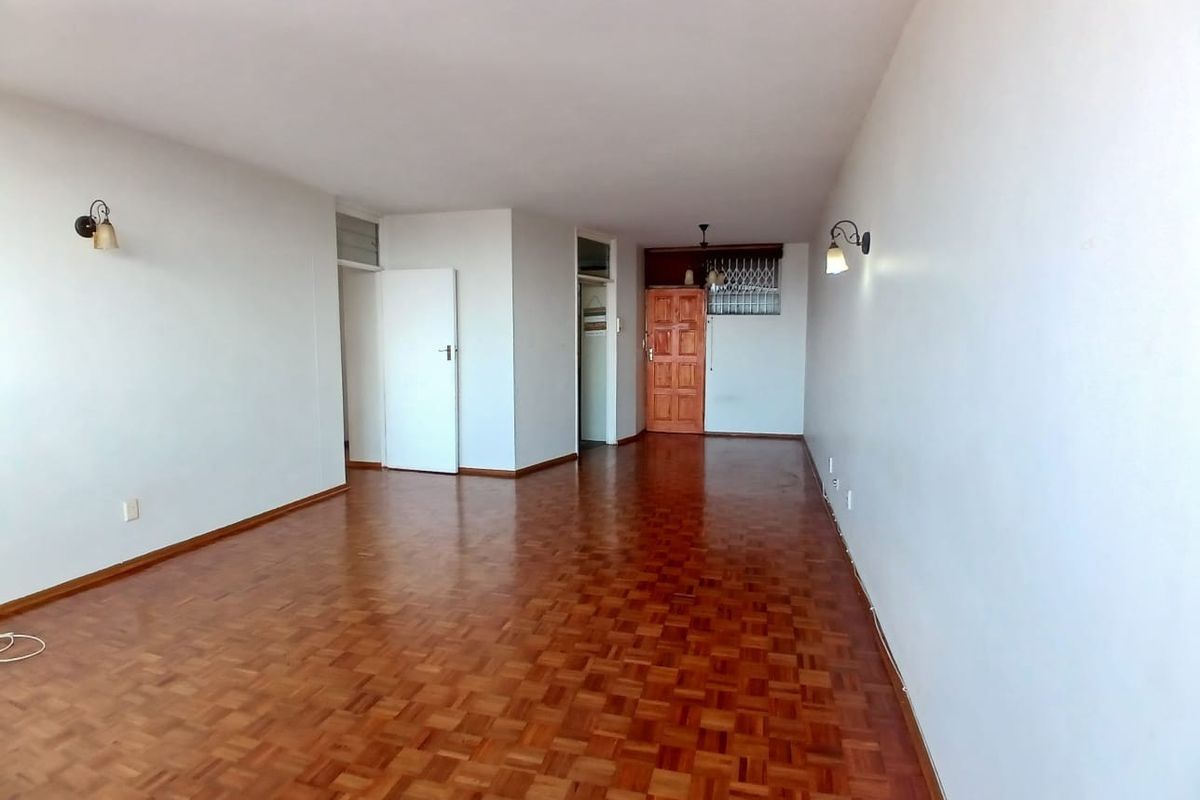 A spacious, empty living area featuring classic parquet flooring, neutral walls, and wall-mounted lighting, providing a versatile blank canvas for interior design.