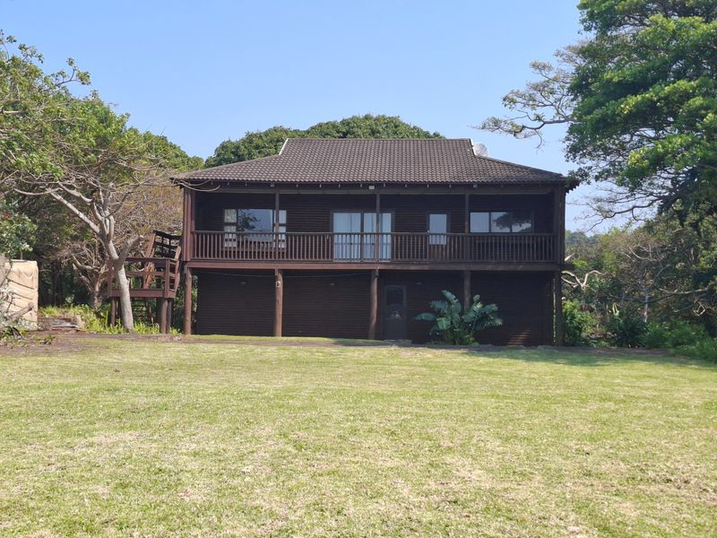 A two-story timber-clad house featuring a raised wooden deck, balcony, and external staircase, set within a spacious garden area.