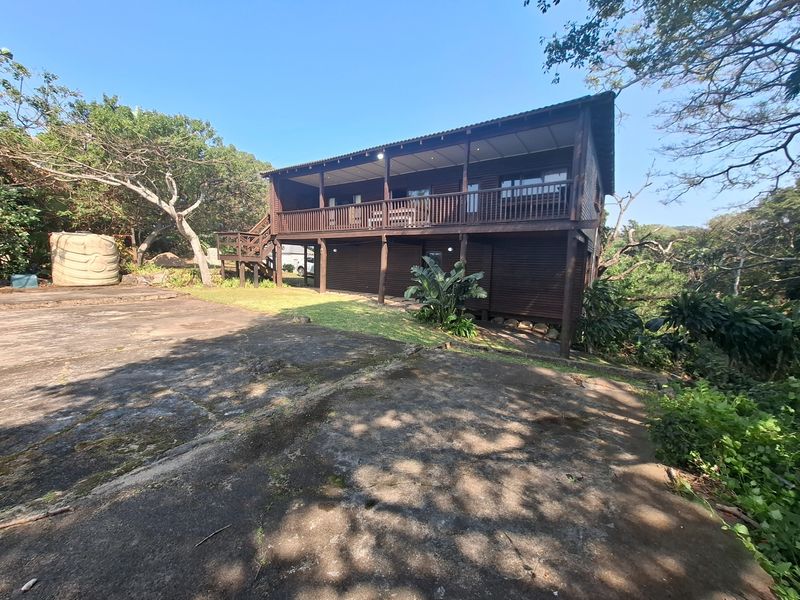 Rear view of an elevated timber-clad house featuring a spacious wooden deck, concrete driveway, and surrounding natural garden setting.