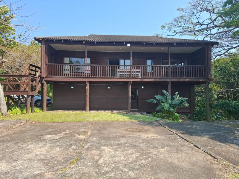 A two-story timber-clad house featuring a spacious raised deck, external staircase, and a large concrete driveway set in a natural, wooded environment.