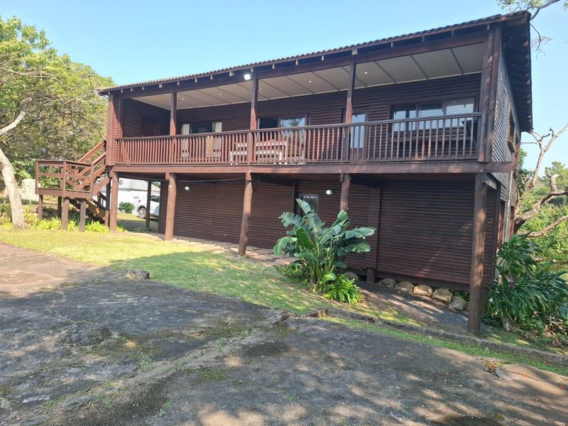 A two-story timber-clad house featuring a spacious raised deck with balustrades, recessed lighting, and an adjacent concrete driveway set in a natural garden environment.