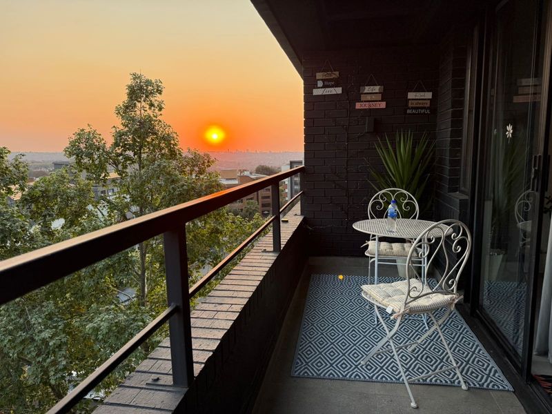 A private balcony featuring a dark brick wall and metal railing, offering an elevated sunset view over the surrounding urban landscape.