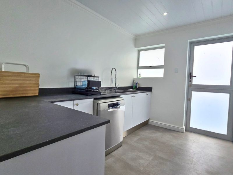A clean and functional scullery featuring modern cabinetry, dark laminate countertops, a stainless steel sink, and direct access to the exterior.