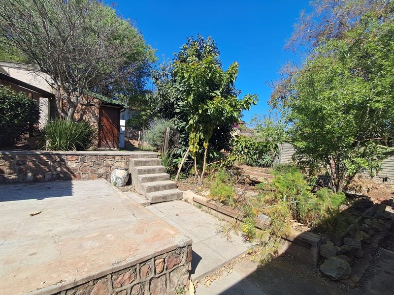 A rear garden area featuring a stone retaining wall, concrete patio, and stairs leading to a side entrance of the house.