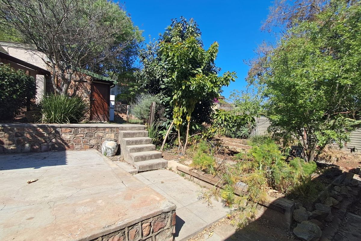A rear garden area featuring a stone retaining wall, concrete patio, and stairs leading to a side entrance of the house.