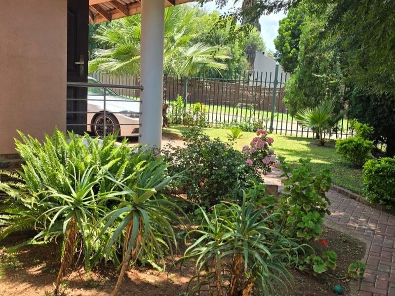 A well-maintained garden area featuring a brick paved pathway, established landscaping, and a covered porch entrance with metal perimeter fencing.