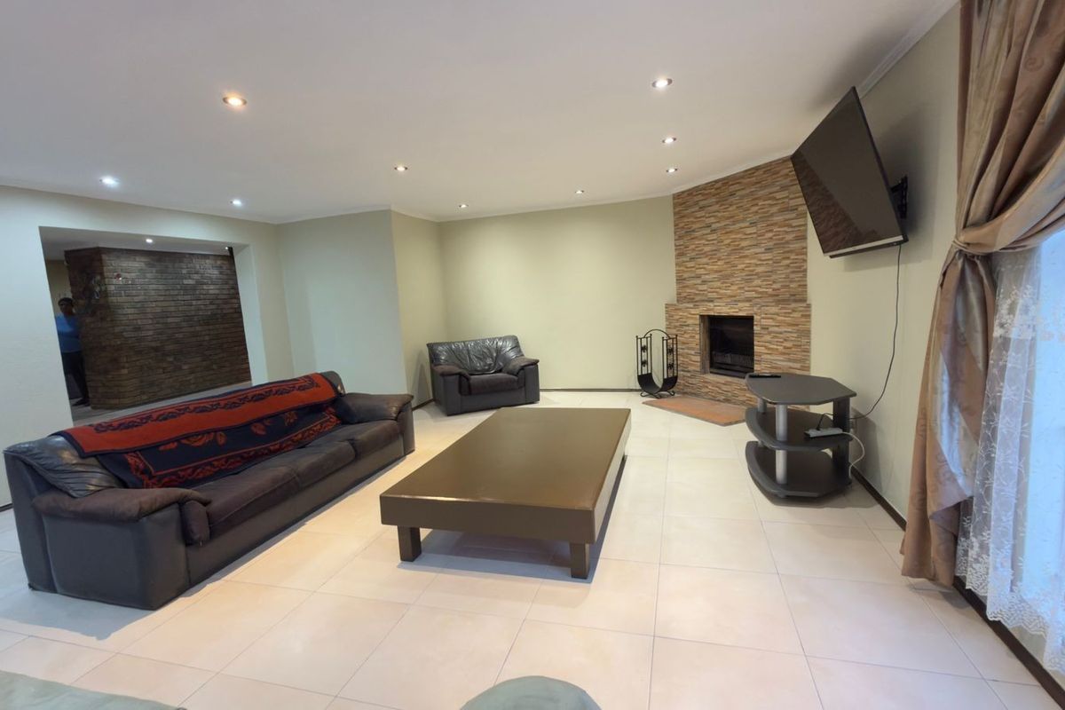 Spacious living room featuring a stone-clad fireplace, recessed downlighting, and neutral tiled flooring, with an adjacent exposed brick feature wall.