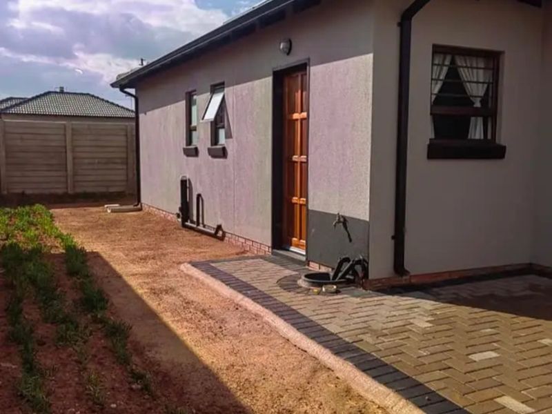 Rear exterior view of the house featuring a paved walkway, solar geyser, and unfinished garden area with boundary walling.