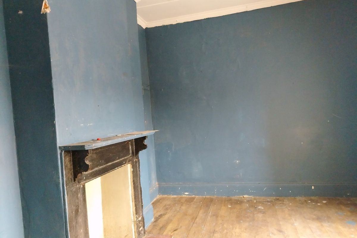 An empty living room featuring original timber flooring and a traditional fireplace mantel, requiring cosmetic refurbishment and wall repairs.