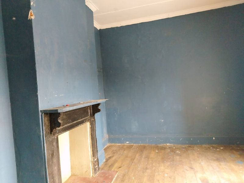 An empty living room featuring original timber flooring and a traditional fireplace mantel, requiring cosmetic refurbishment and wall repairs.