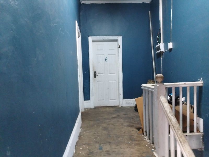 A narrow commercial hallway featuring dark blue walls, a white panel door, and a wooden staircase balustrade, currently in need of cosmetic refurbishment.