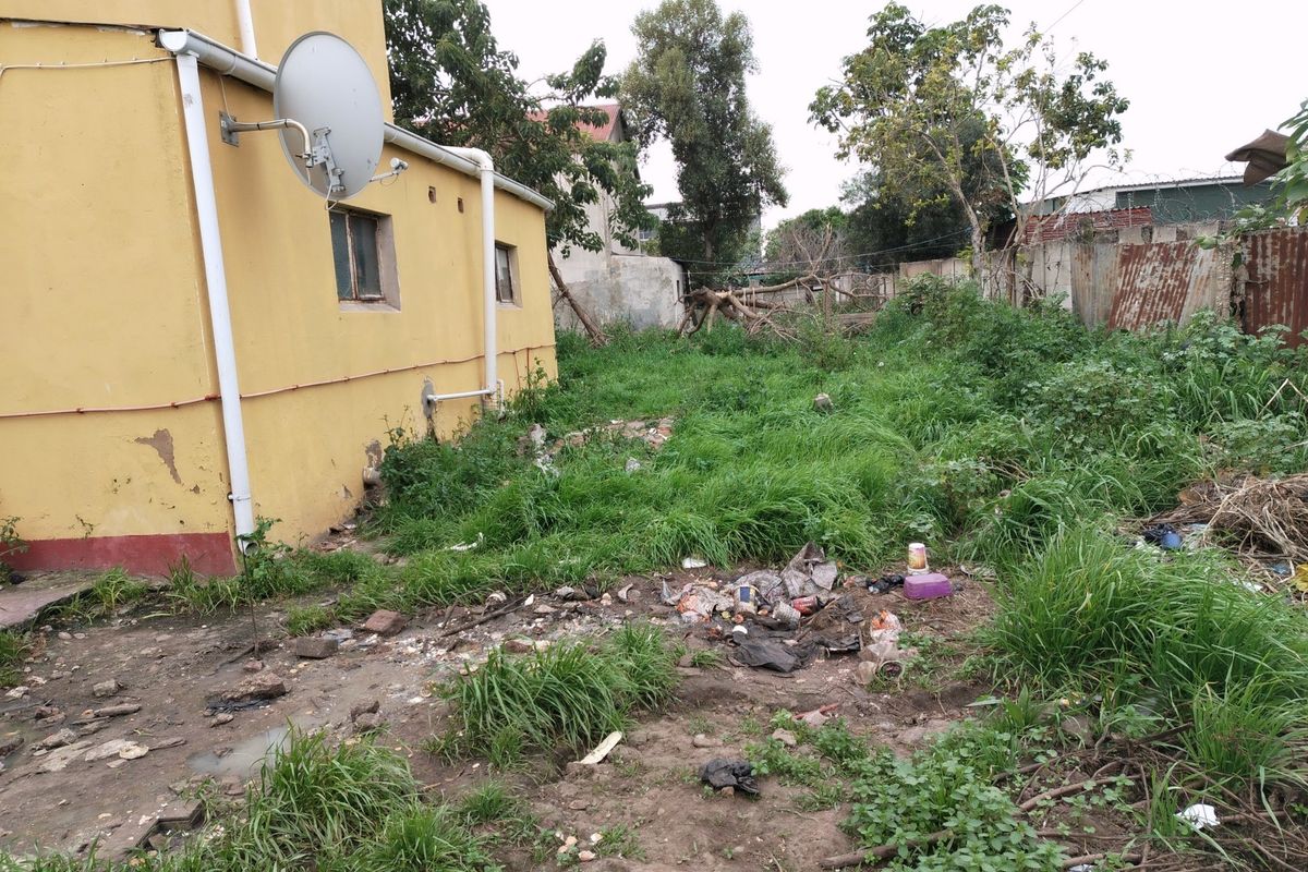 Rear exterior view of the commercial property showing the building facade, downpipe, satellite dish, and an unmaintained yard area with overgrown grass and debris.