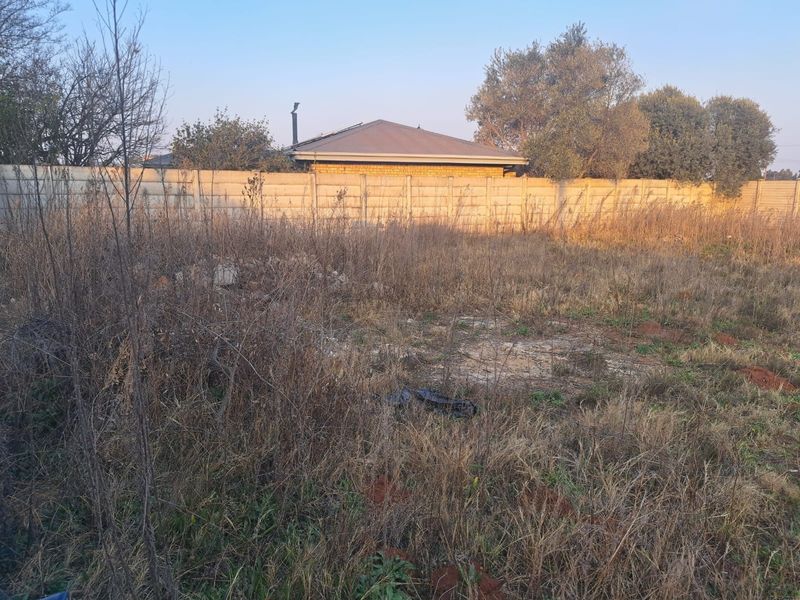 A view of a vacant commercial plot featuring overgrown vegetation and a precast concrete boundary wall at the rear.