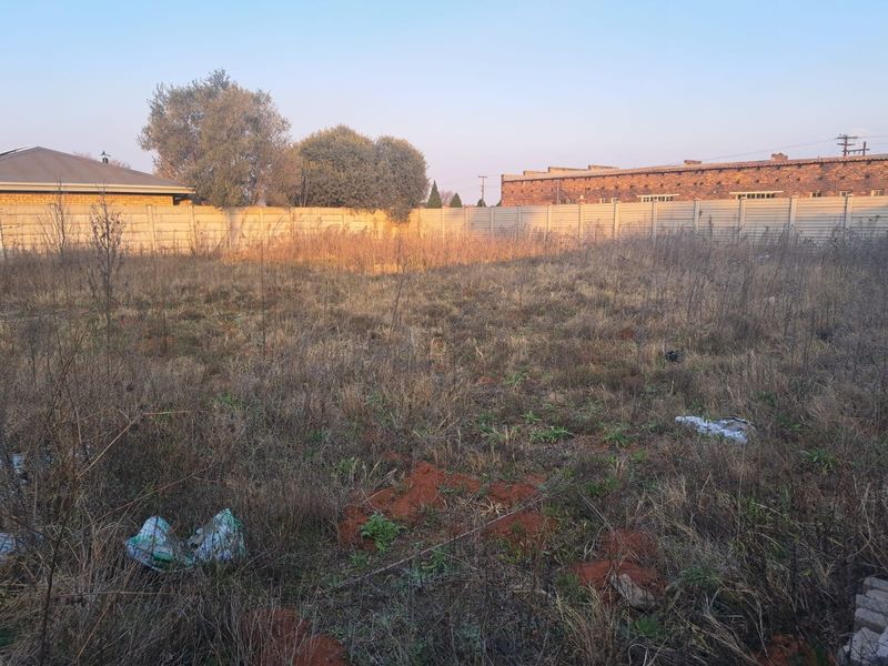 A view of a vacant commercial plot featuring dry, overgrown vegetation and enclosed by a precast concrete boundary wall.