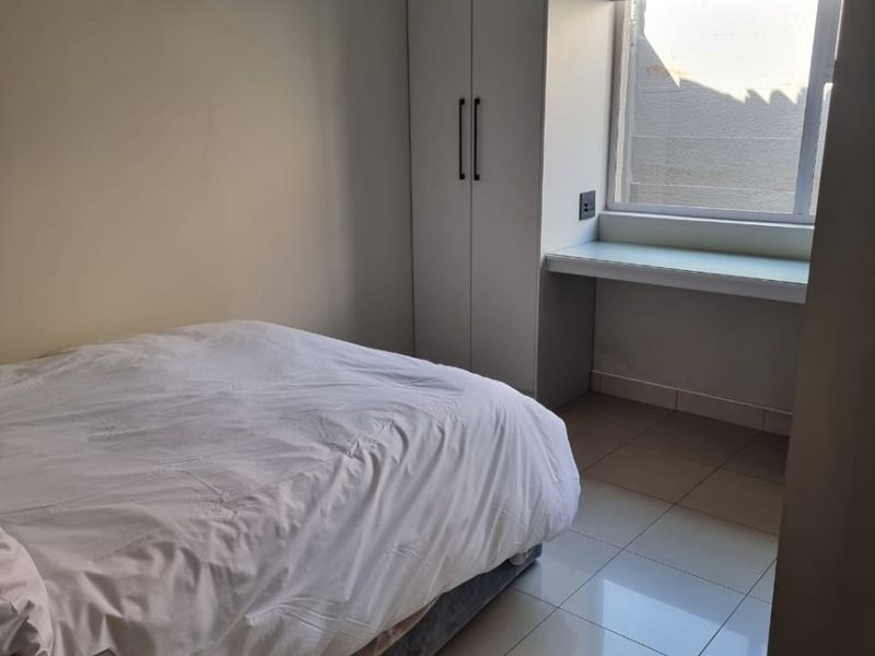 A clean, modern bedroom featuring neutral walls, tiled flooring, a built-in wardrobe, and a convenient integrated desk space by the window.