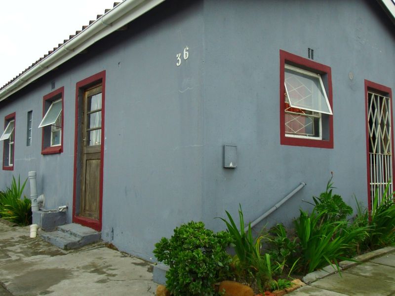 Side and rear view of a residential house featuring grey exterior walls, red-trimmed windows with security gates, and concrete paved surrounds.