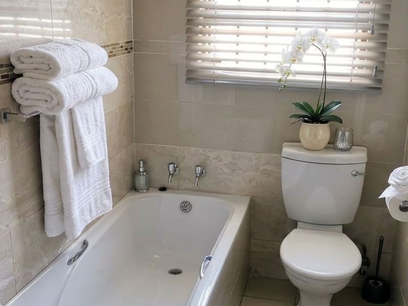 A well-maintained bathroom featuring neutral wall tiles, a built-in bathtub, and a standard toilet with window blinds for privacy.