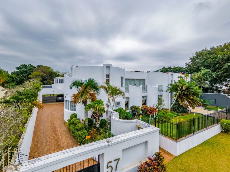 A modern, white multi-level residential property featuring a curved architectural design, paved driveway, carport, and landscaped front garden with perimeter fencing.