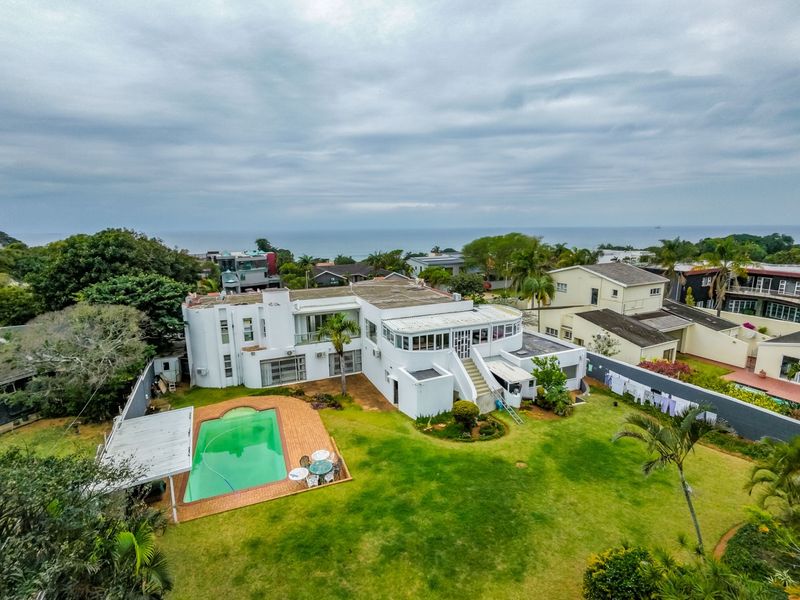 Aerial view of a large residential property featuring a swimming pool, expansive lawn, and multi-level exterior structure with ocean views in the distance.