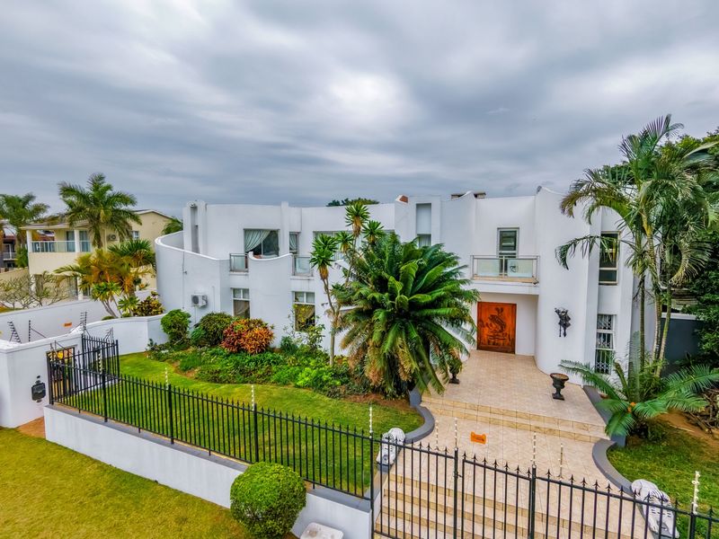A striking modern multi-story residence featuring a curved white facade, tiled entrance patio, glass-railed balconies, and a well-maintained landscaped front garden.