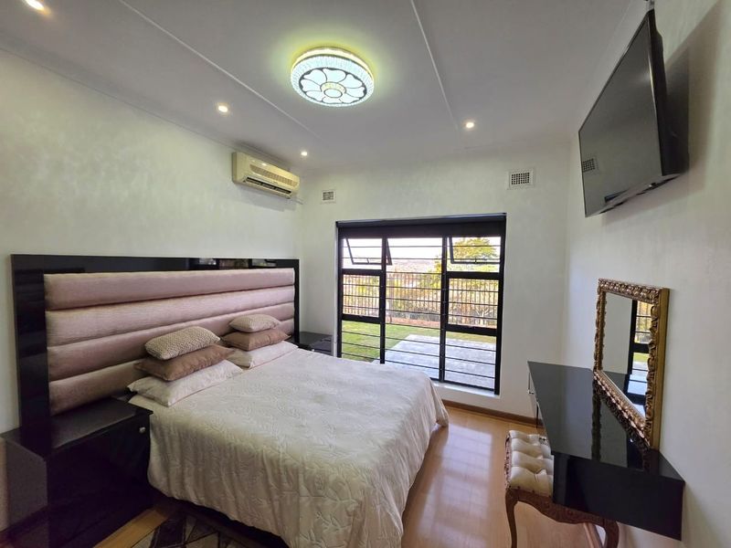 A well-maintained bedroom featuring laminate flooring, an integrated headboard, air conditioning, and large windows with security bars overlooking the garden.