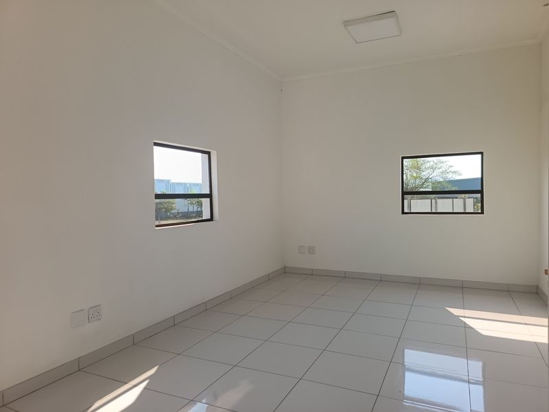 A bright, empty office space featuring clean white walls, polished tiled flooring, and large aluminium-framed windows providing natural light.